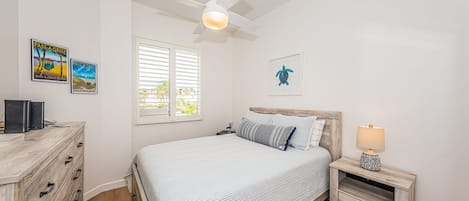 Un dormitorio con cama, mesita de noche, escritorio, ventilador de techo y dos cuadros colgados.