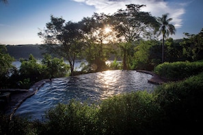 Una piscina circular con borde de piedra, rodeada de abundante follaje y árboles, con el sol poniéndose al fondo.