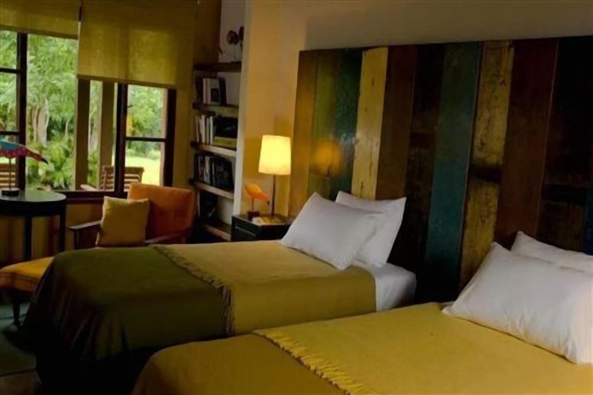 Ein Hotelzimmer mit zwei Betten, einem kleinen Tisch mit Stuhl, einem Bücherregal und einem Fenster mit Blick auf Grünflächen.
