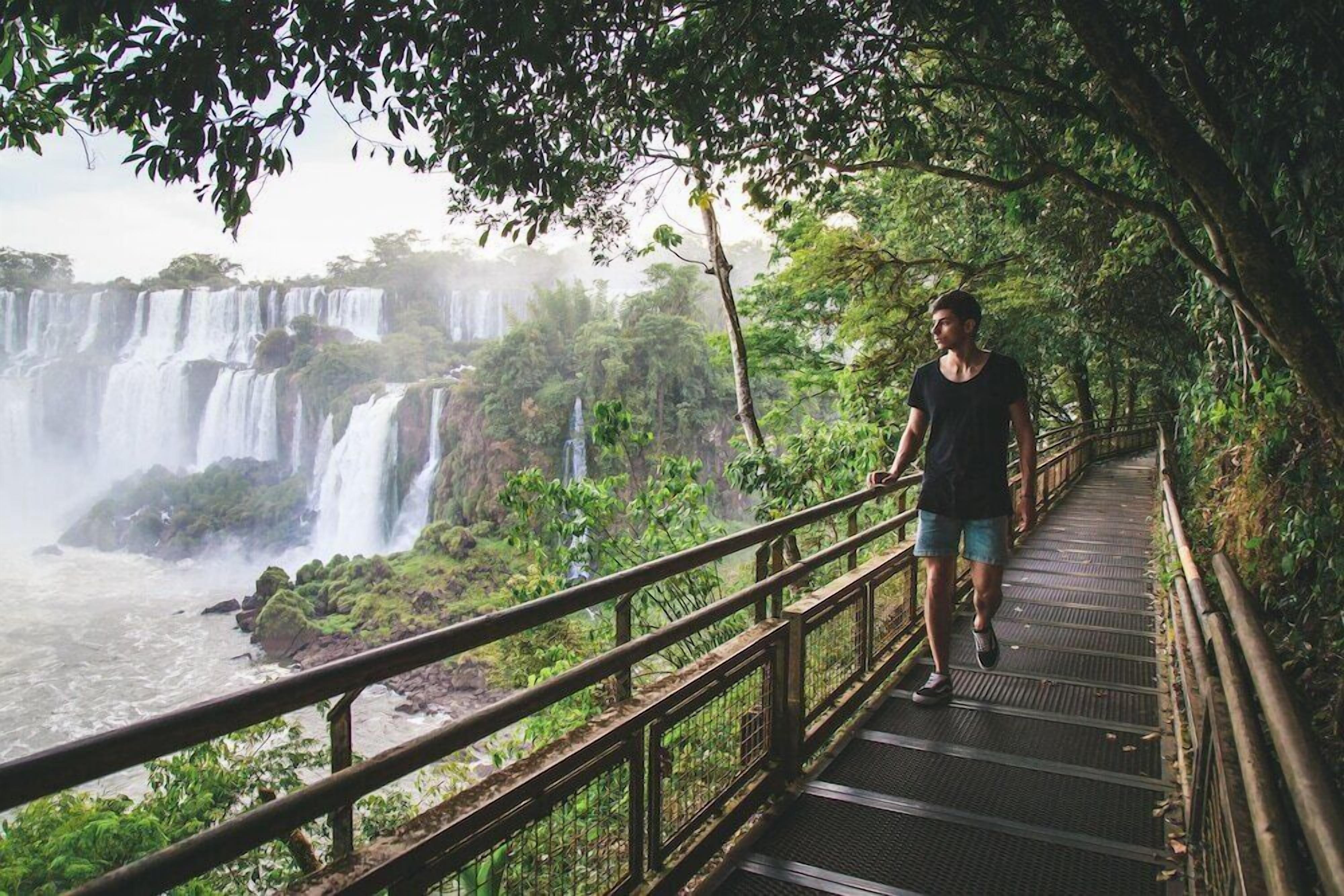Una persona che cammina su un ponte di legno con ringhiere metalliche, con vista su una serie di cascate e vegetazione rigogliosa.