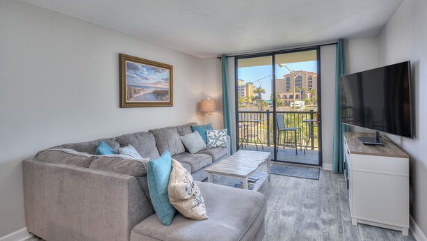 Un salon moderne avec un canapé d’angle gris, un téléviseur à écran plat et un balcon offrant une vue sur les immeubles.