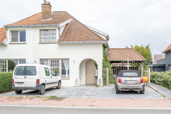 A white two-story house with a tiled roof, a driveway, and two parked cars. - Diane's Cottage (Knokke-Heist)