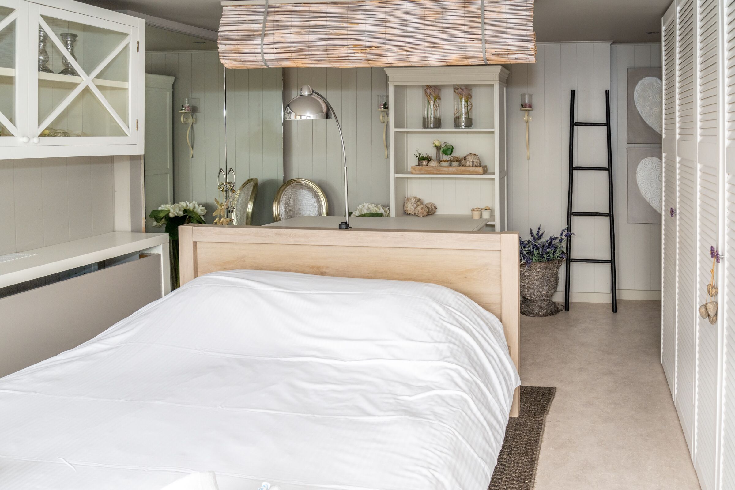 Een slaapkamer met een houten bed, een witte kledingkast, een spiegel en een plank met decoratieve objecten.
