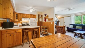 Une cuisine avec des armoires en bois, une cuisinière et un réfrigérateur. On y trouve une table à manger et des tabourets de bar. Une chaise en bois est visible au fond.