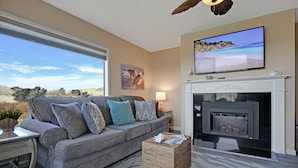 A living room with a grey sofa, a fireplace, a television, and a large window overlooking a natural landscape. - Remote Oceanfront Retreat Between Santa Cruz & Monterey (Watsonville)