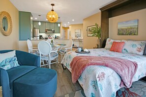 A cozy hotel room with a bed, a dining table, and chairs. There is a kitchen area in the background.
