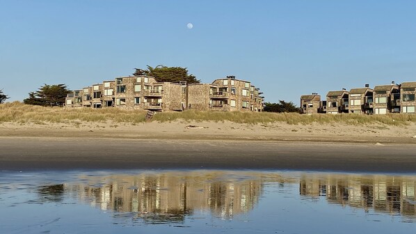 A beachfront property with multiple buildings, a sandy beach, and a calm body of water reflecting the structures.