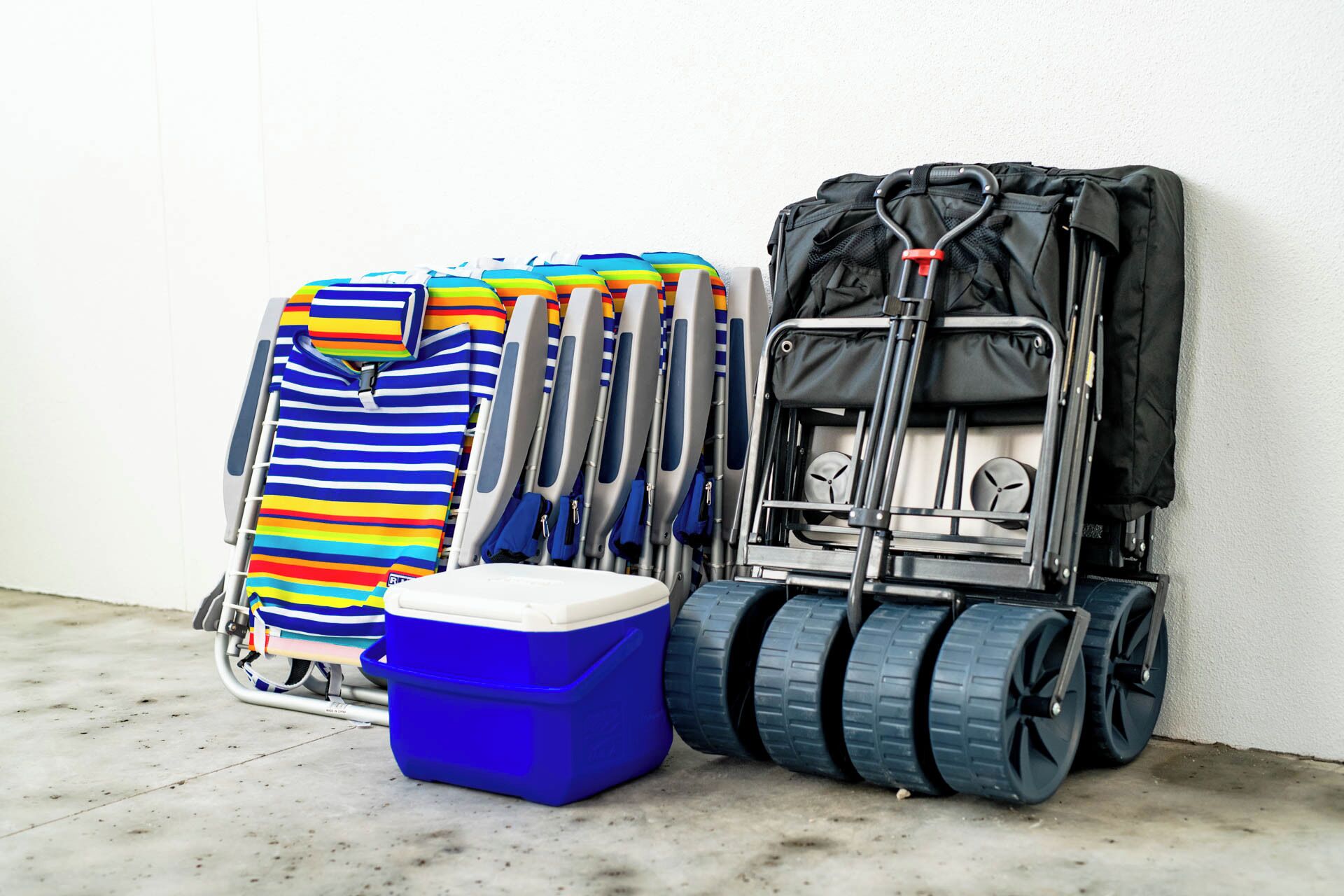 A luggage cart with a blue cooler, a striped beach towel, and a black suitcase against a white wall.