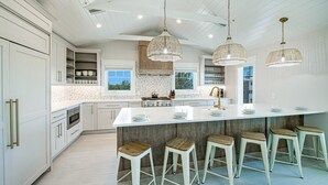 A modern kitchen with white cabinetry, a large island with stools, pendant lights, and a view of the outdoors through windows. - Nearly Beachfront, One house from the beach! Pool and Game Room - Ocean Overlook (Siesta Key)