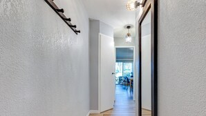 Un corridoio con pavimento in legno, pareti bianche, uno specchio con cornice nera e un lampadario.
