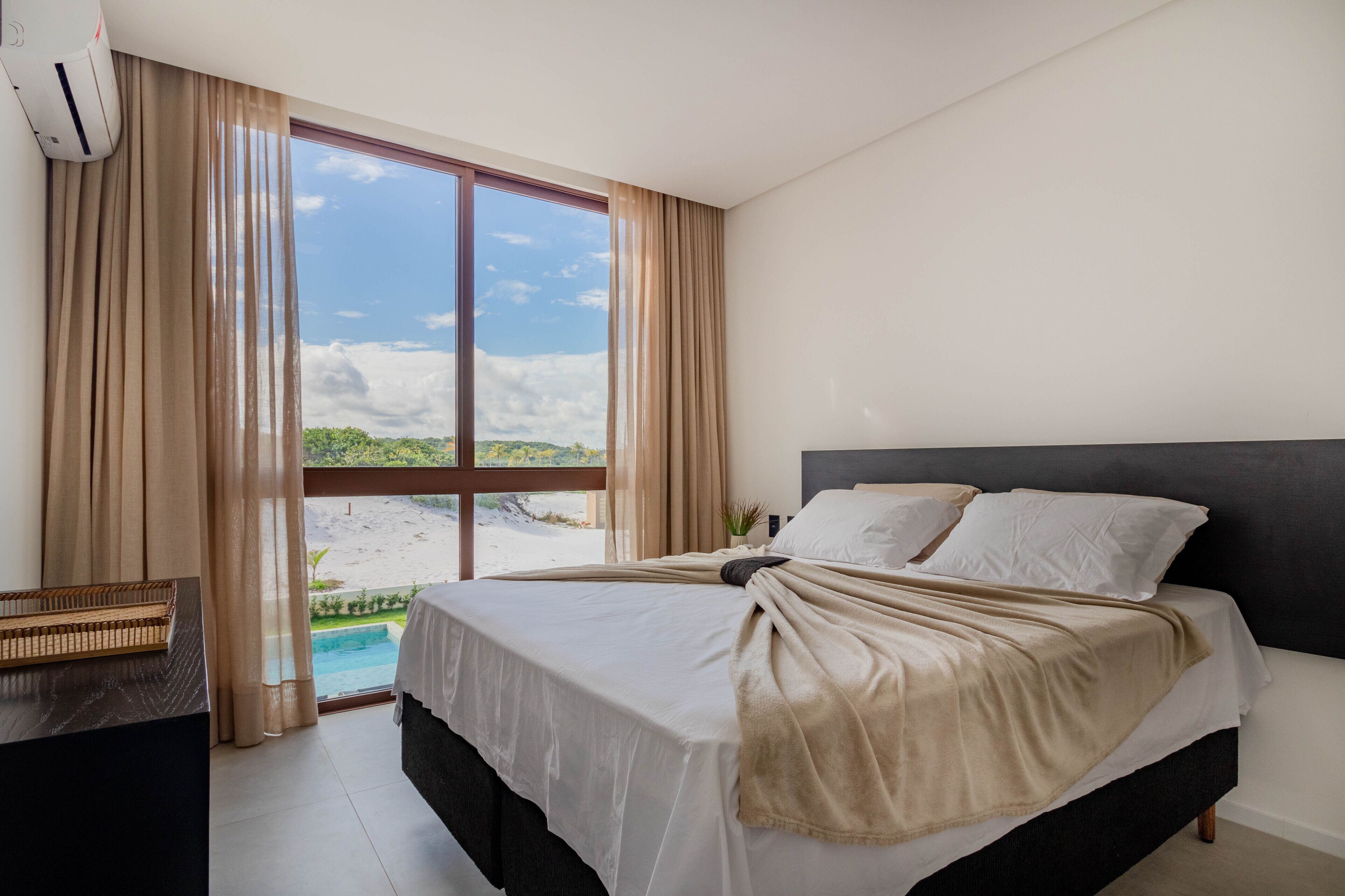 Quarto de hotel com uma cama grande, uma mesa de cabeceira e vista para uma praia e piscina.