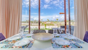 Une table à manger dressée pour quatre personnes, agrémentée d’un grand saladier décoratif, d’assiettes et de couverts, avec vue sur une plage et des palmiers.