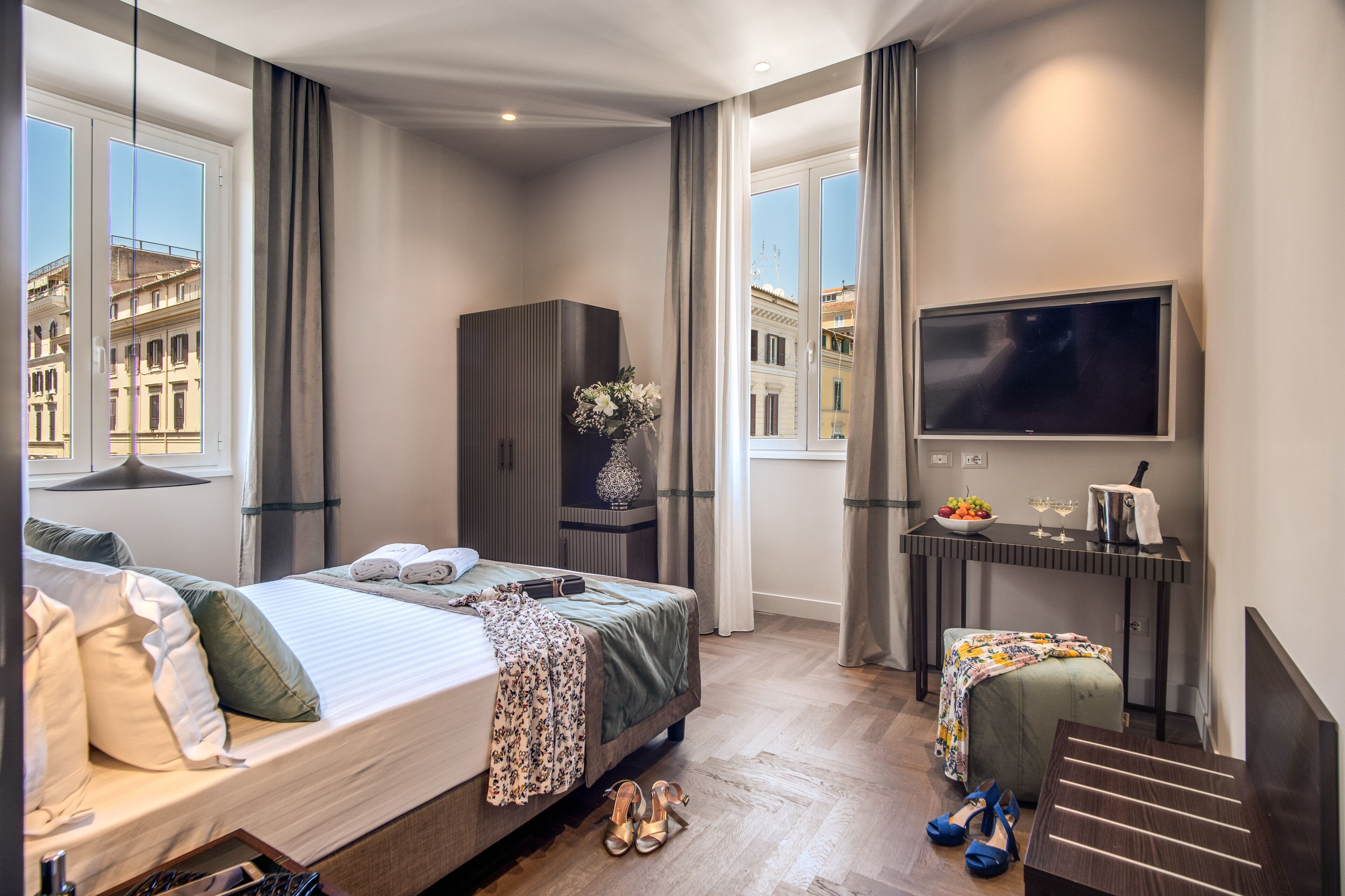 A hotel room with a bed, a desk with a TV, a wardrobe, and a view of buildings.