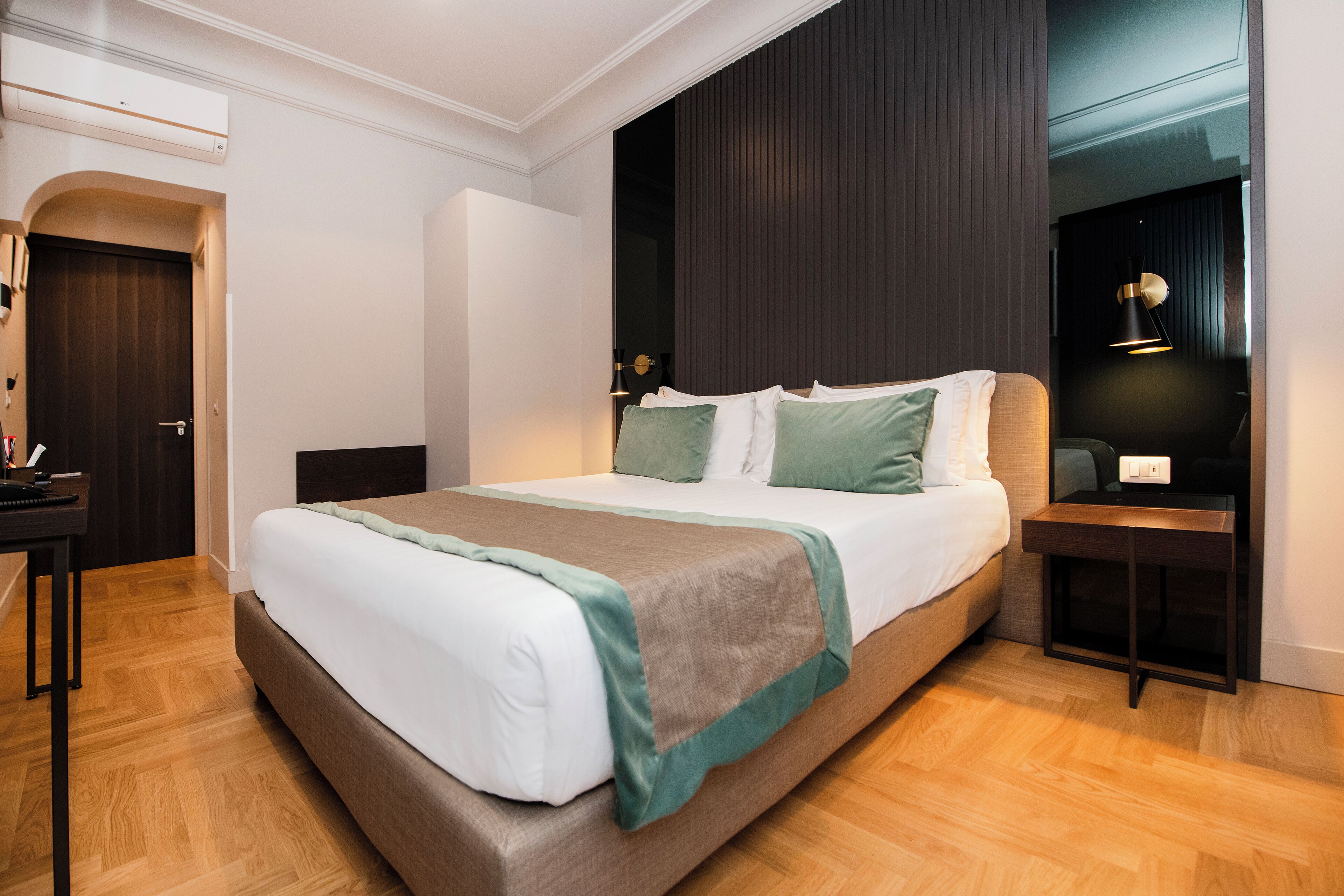 A modern hotel room with a large bed, a bedside table, a desk, and a wardrobe.
