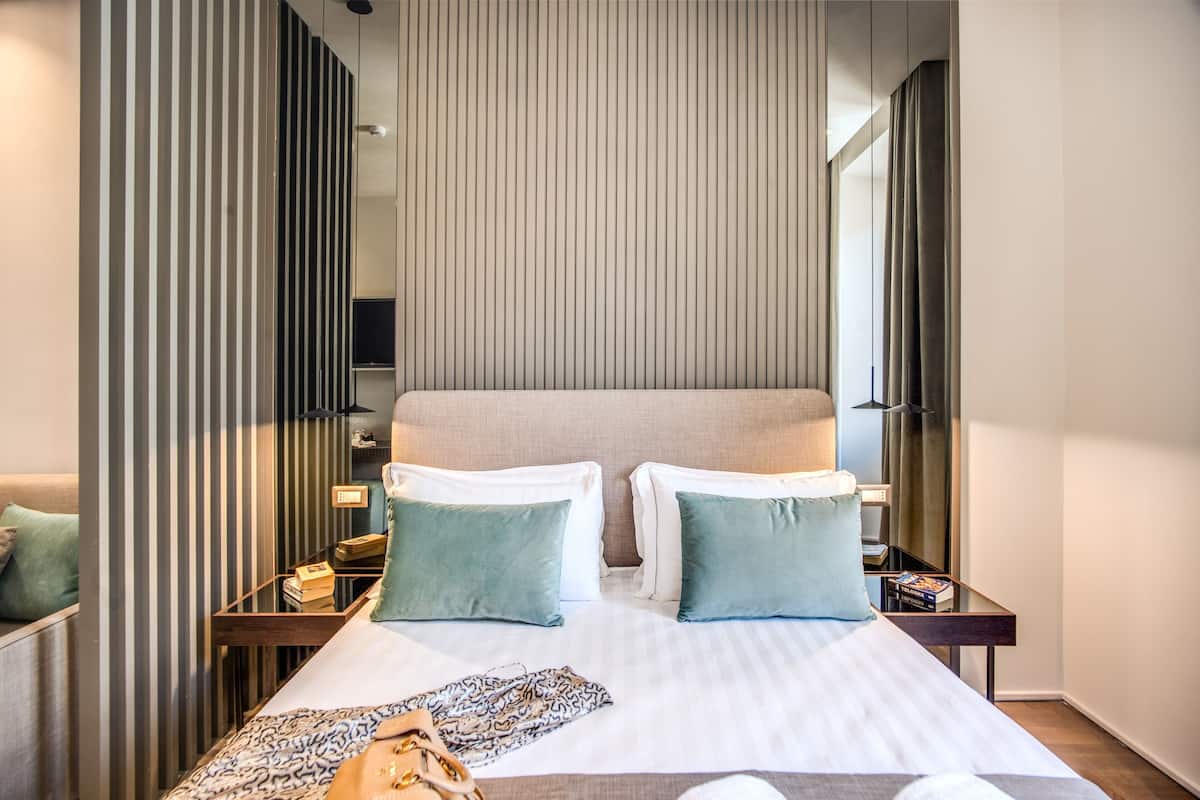 Hotellihuone, jossa on suuri sänky, kaksi yöpöytää, yölamppu, peili ja ikkuna, jossa on verhot.