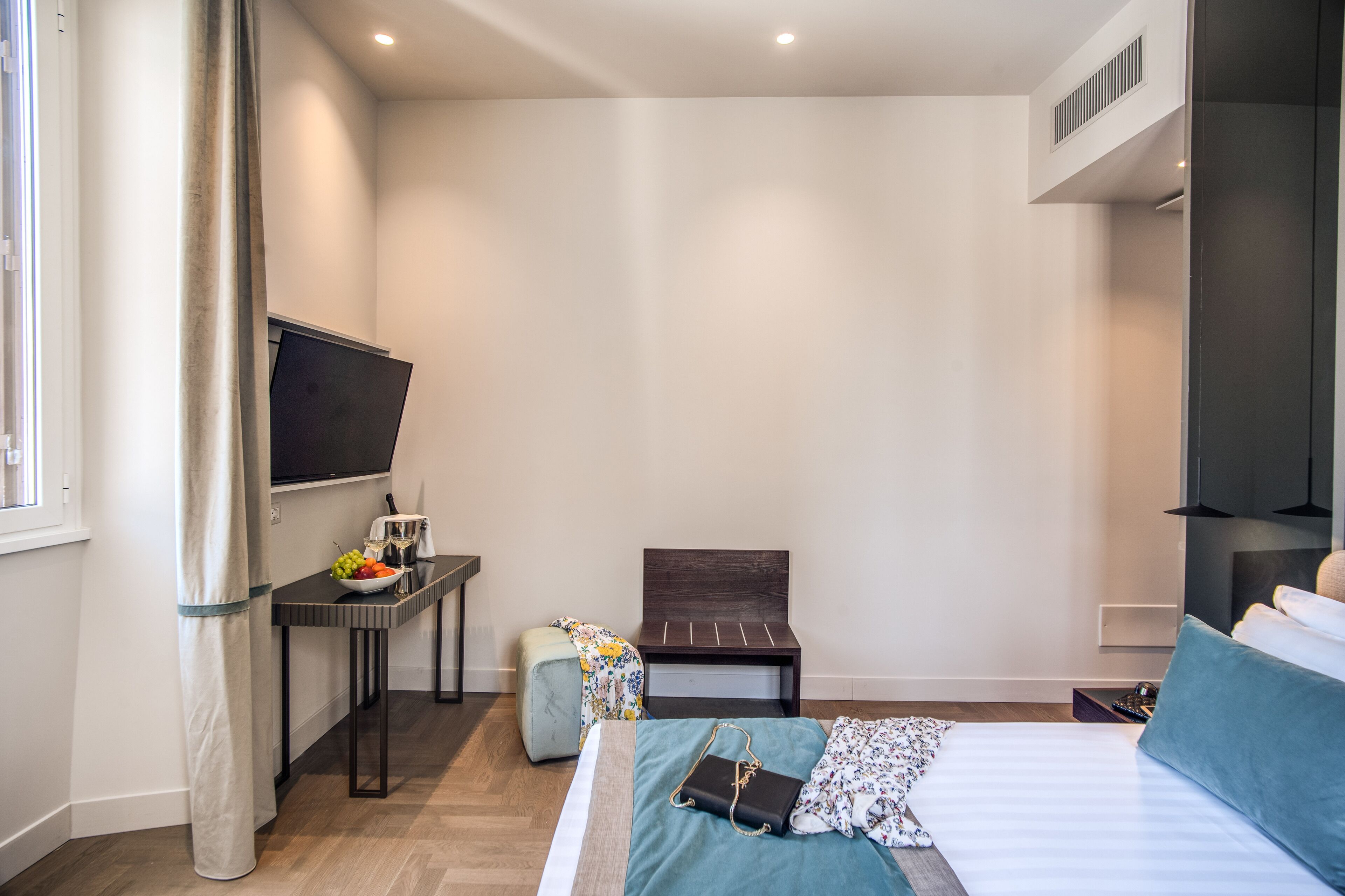 A modern hotel room with a bed, a desk with a chair, a small table with fruits, a TV, and a window with curtains.