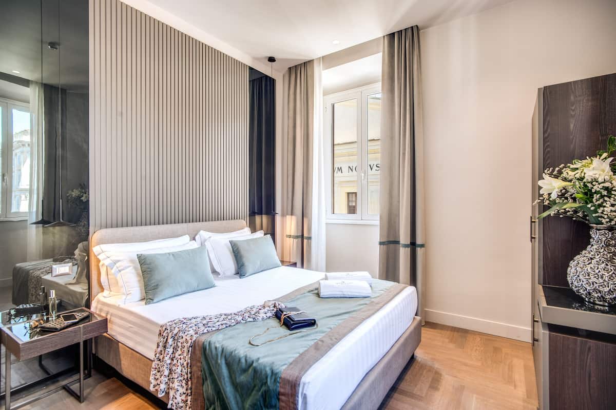 Moderni hotellihuone, jossa on suuri sänky, yöpöytä lampun kanssa, peili ja ikkunasta avautuva näkymä kaupunkiin.