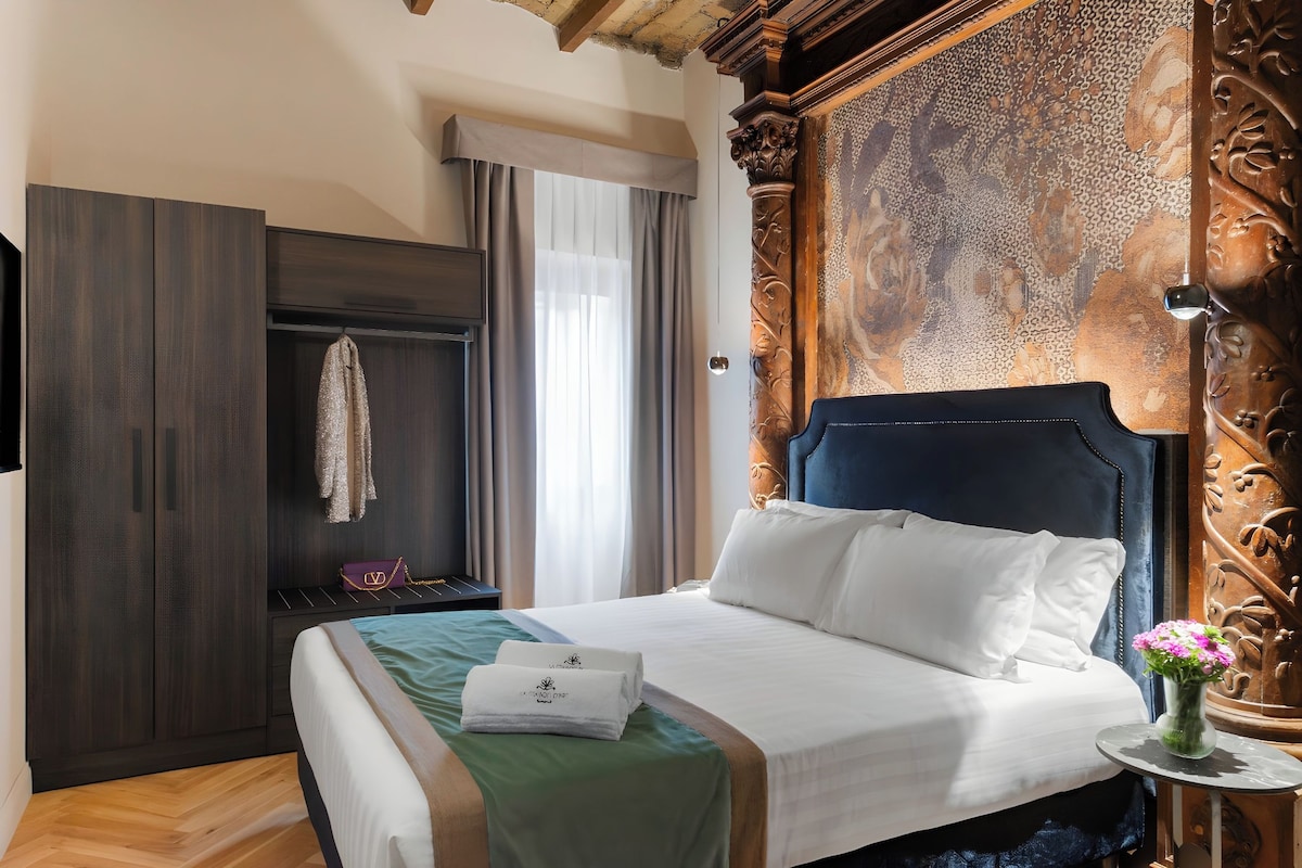A hotel room with a large bed, a wardrobe, a vanity with a mirror, and a small table with a vase of flowers.