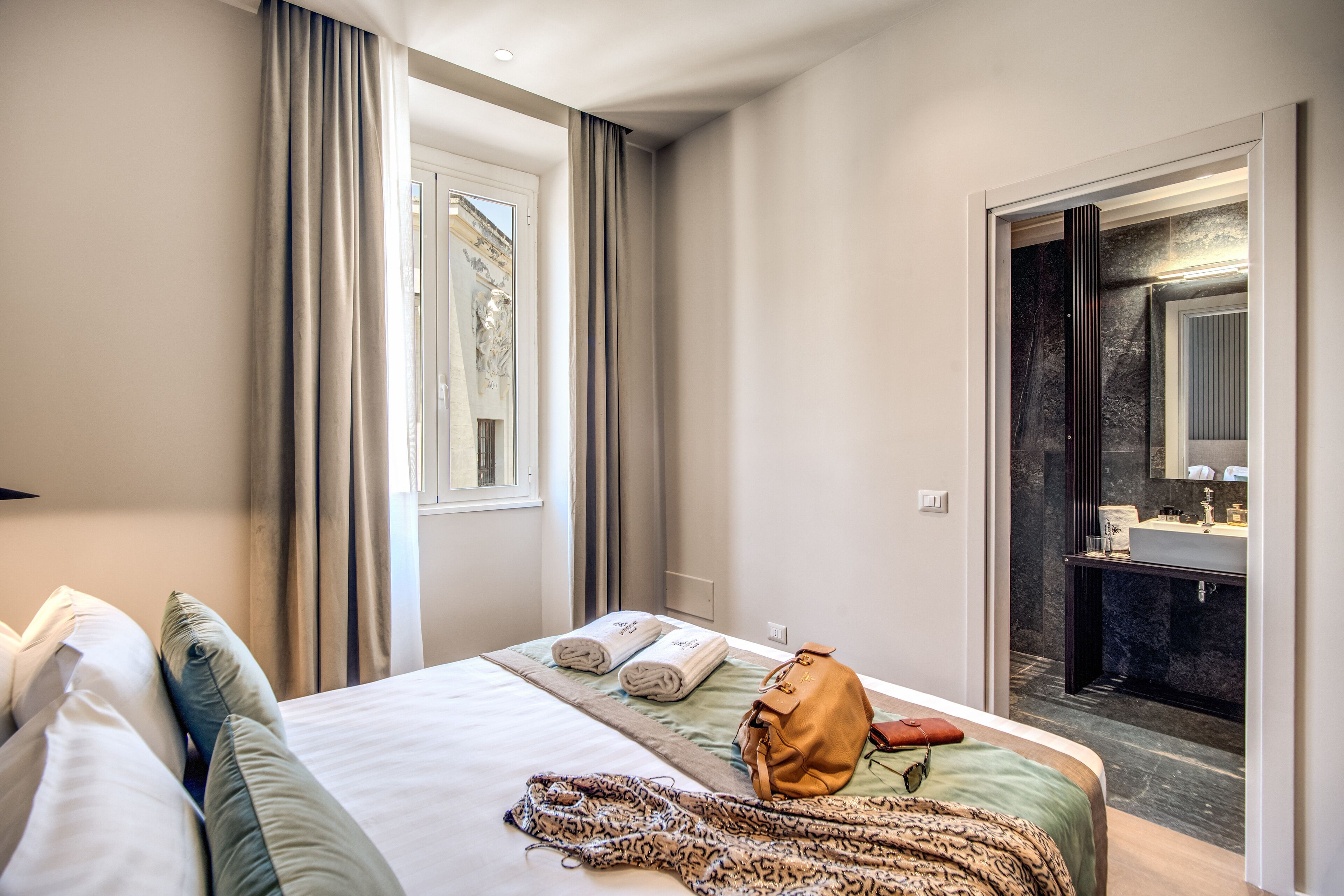 A hotel room with a bed, a bathroom visible through an open door, and a window with curtains.