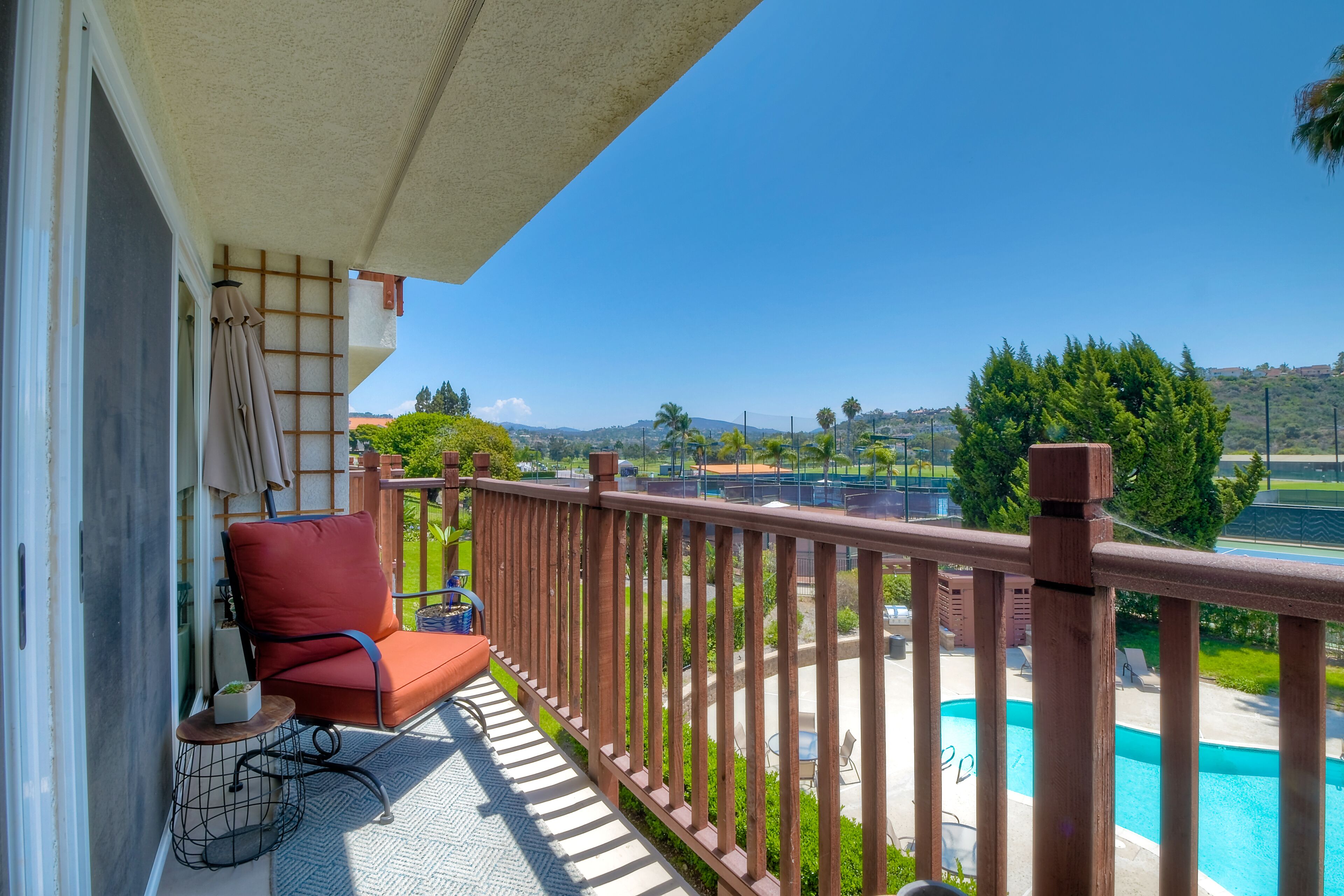 A balcony with a red chair, a small table with a plant, and a view of a swimming pool and greenery.