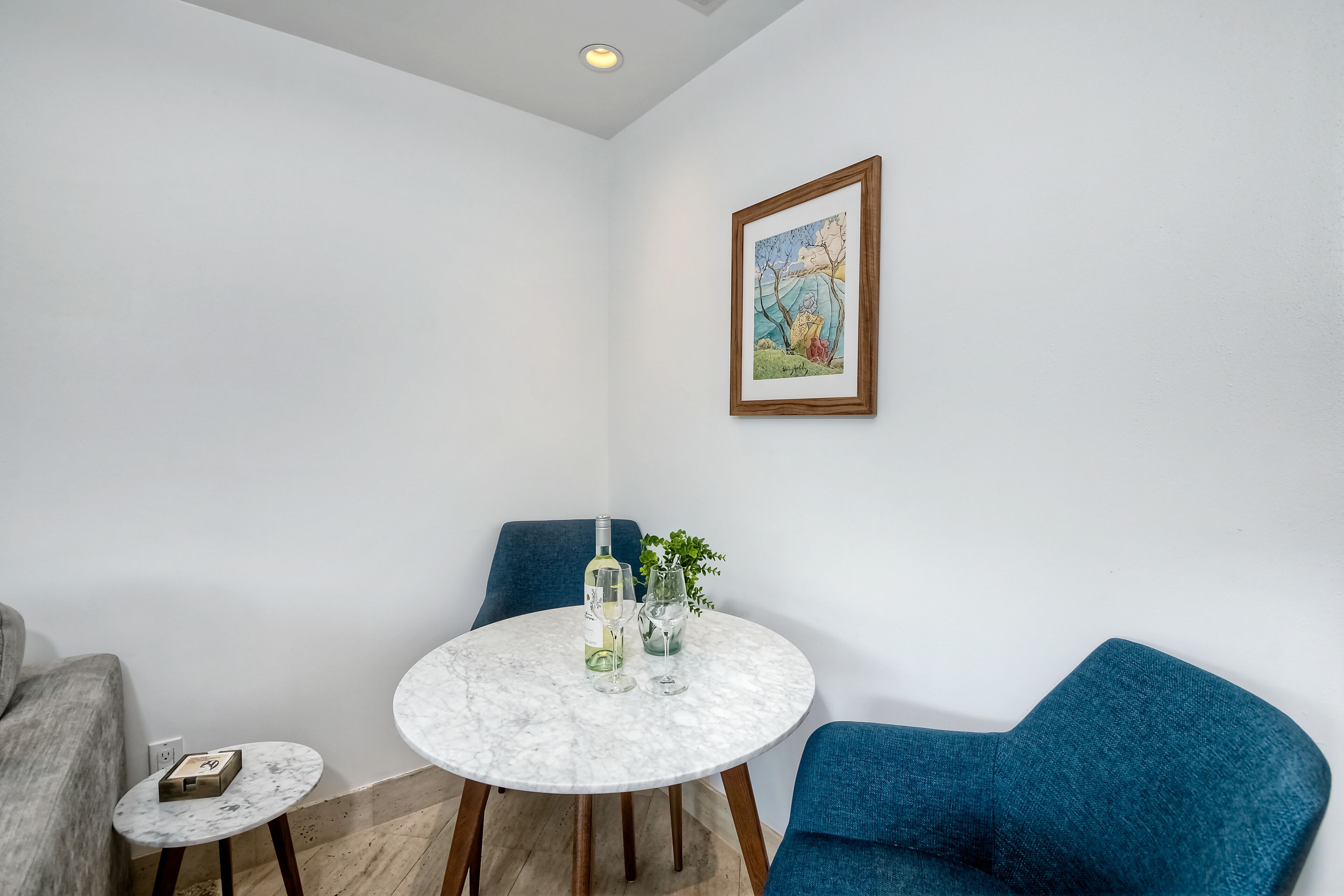 Une pièce moderne avec une table ronde en marbre, deux chaises bleues, une petite table d’appoint et un tableau encadré accroché au mur.