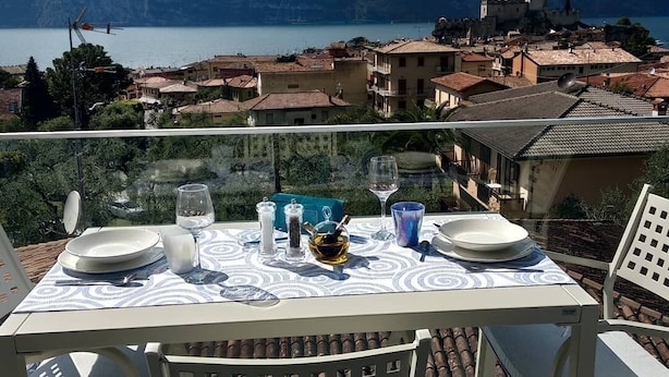 A table set for two with a view of a lake and mountains in the background. - BLUE SKY SAPPHIRE APP.  LAKE VIEW CIR:023045-LOC-00628 CIN: EN023045C2HTNJMTGL (Malcesine)