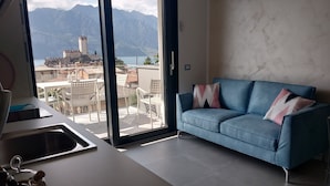 A modern living room with a blue sofa, a kitchen area with a sink and a bottle, and a large sliding glass door leading to a balcony with a mountain view. - BLUE SKY SAPPHIRE APP.  LAKE VIEW CIR:023045-LOC-00628 CIN: EN023045C2HTNJMTGL (Malcesine)