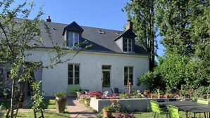 Ein weißes Haus mit dunklem Dach, einer Veranda mit Stühlen und einem Garten mit Blumentöpfen und Blumen.