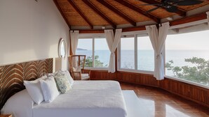 Un dormitorio con una cama grande, vigas de madera en el techo, un ventilador de techo y vista al mar.