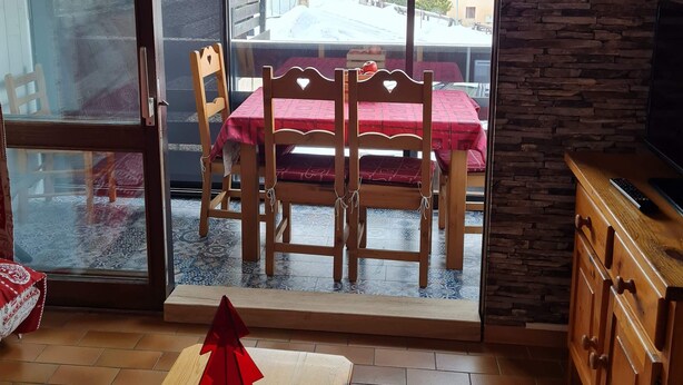 A room with a wooden dining table and chairs, a red tablecloth, a wooden cabinet, and a view of a snowy landscape through a glass door. - Chaleureux Appartement (Ancelle)