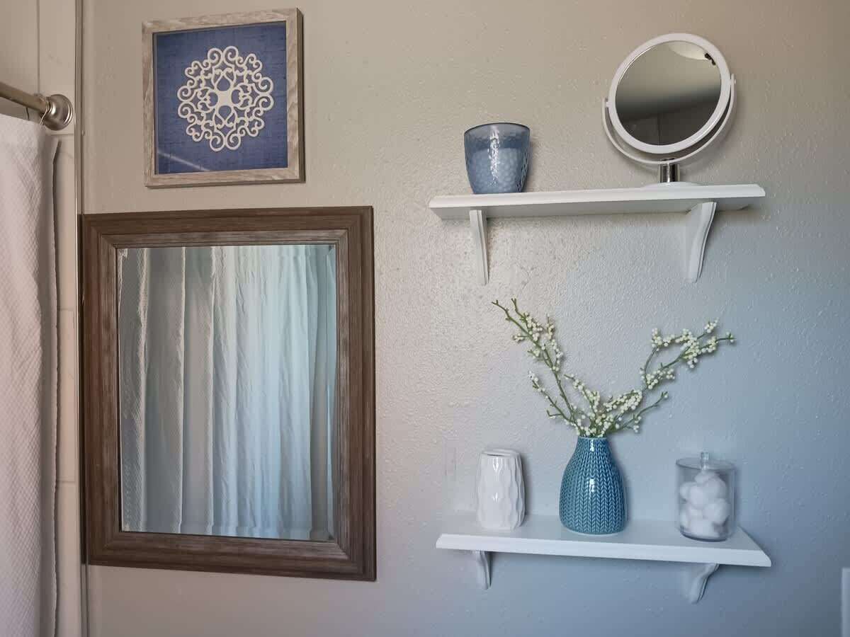 A bathroom with a wooden-framed window, a decorative wall piece, a mirror, and a shelf with a vase and candle.