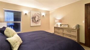 Ein Schlafzimmer mit Bett, Kommode, Wandbild und Fenster mit Jalousien.