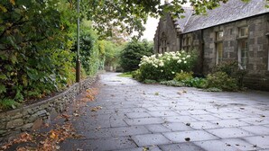 Ein gepflasterter Weg führt zu einem Steinhaus mit traditioneller Gestaltung, umgeben von üppigem Grün und weißen Blüten.