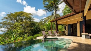 A swimming pool with clear water, surrounded by lush greenery and a covered patio area with lounge chairs.