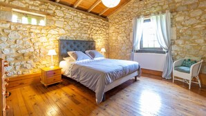 A bedroom with a stone wall, wooden floor, a bed with a headboard, a nightstand with a lamp, a window with curtains, and a chair.