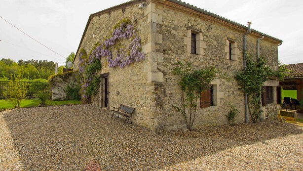 A stone building with a gravel driveway, a bench, and a pergola with purple vines.