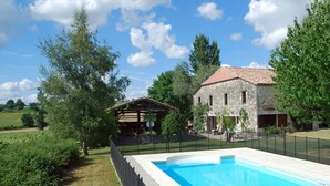 A stone house with a red-tiled roof, a swimming pool, and a fenced garden area.