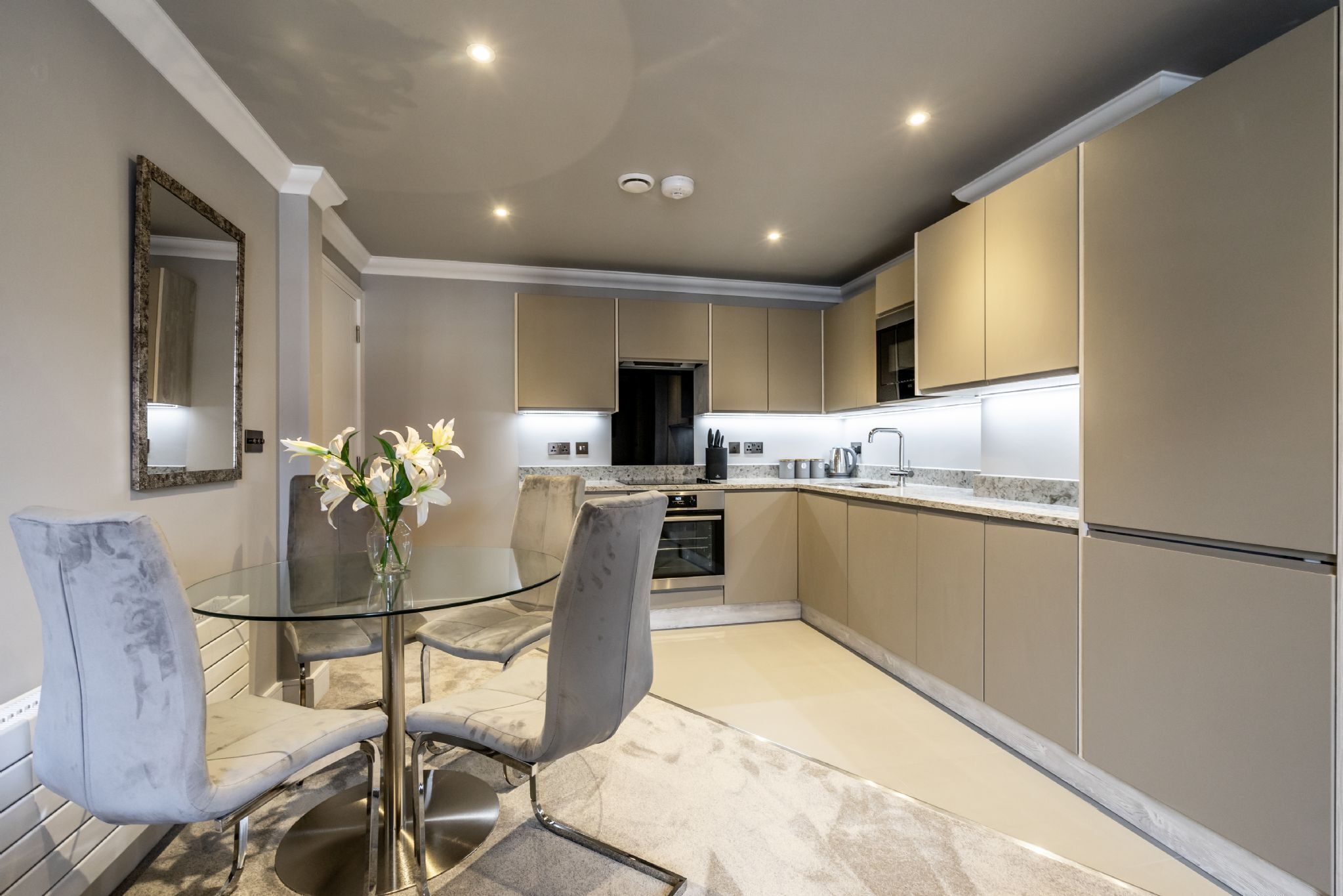 Une cuisine moderne avec une table à manger en verre, des chaises à revêtement gris et des meubles de rangement de couleur beige.