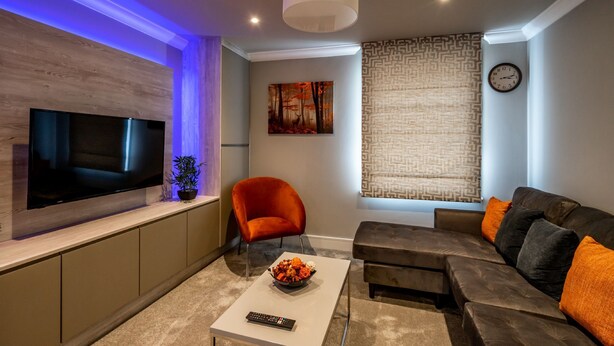A modern living room with a flat-screen TV, a red chair, a sofa with orange cushions, a coffee table with a bowl of fruit, and a patterned curtain. - Bootham Urban Space -  a family break that sleeps 4 guests  in 2 bedrooms (York)