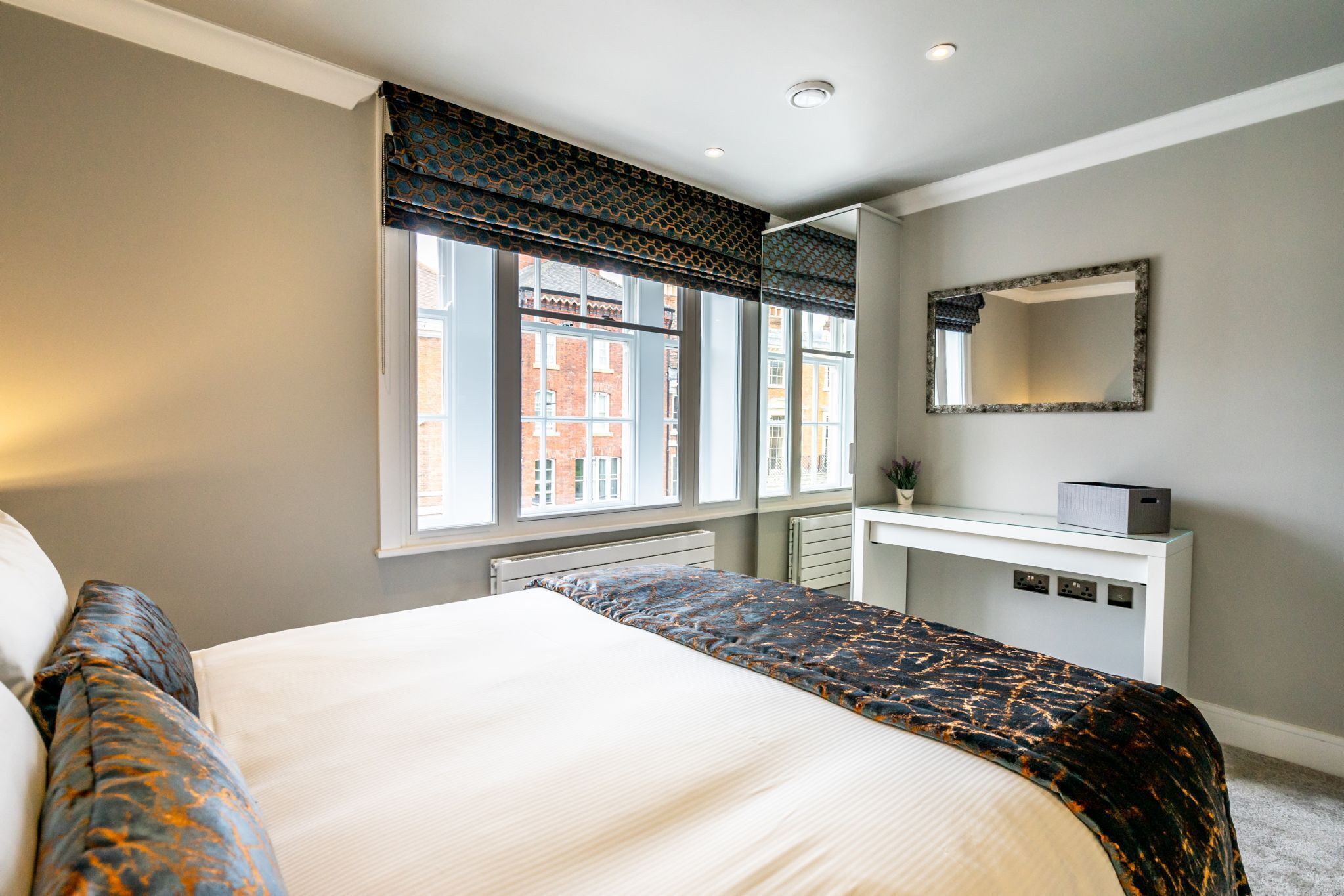 Une chambre à coucher avec un grand lit, une table de chevet, un miroir et une fenêtre donnant sur des immeubles.