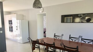 Un coin repas avec une table en bois et six chaises, une cuisine à l’arrière-plan et une horloge murale.
