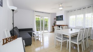 A modern living room with a grey sofa, a white dining table with six chairs, a ceiling fan, and a TV mounted on the wall. - Villa  ADELFA (Salou)