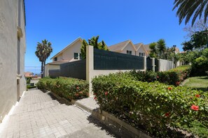 A residential area with a paved pathway, a gated entrance, and lush greenery including red flowers and palm trees.