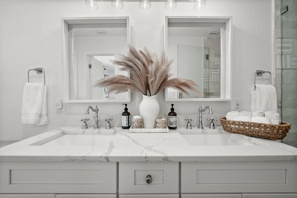 A modern bathroom with a large white marble sink, two mirrors, a vase with pampas grass, and a basket of towels.