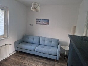 A room with a light blue sofa, a small round table, a radiator, a window with blinds, and a wall-mounted picture. - Idyllic apartment on the shores of Lake Krüpelsee (Königs Wusterhausen)
