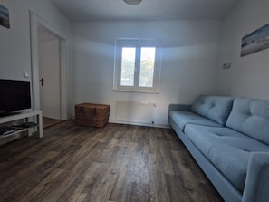 Una sala de estar con televisor, un sofá azul, una cesta de mimbre y una ventana con persianas.