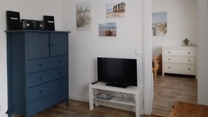 Ett rum med ett blått skåp, ett vitt TV-bord och en vit byrå. Det finns konstverk på väggarna och en flätad stol.