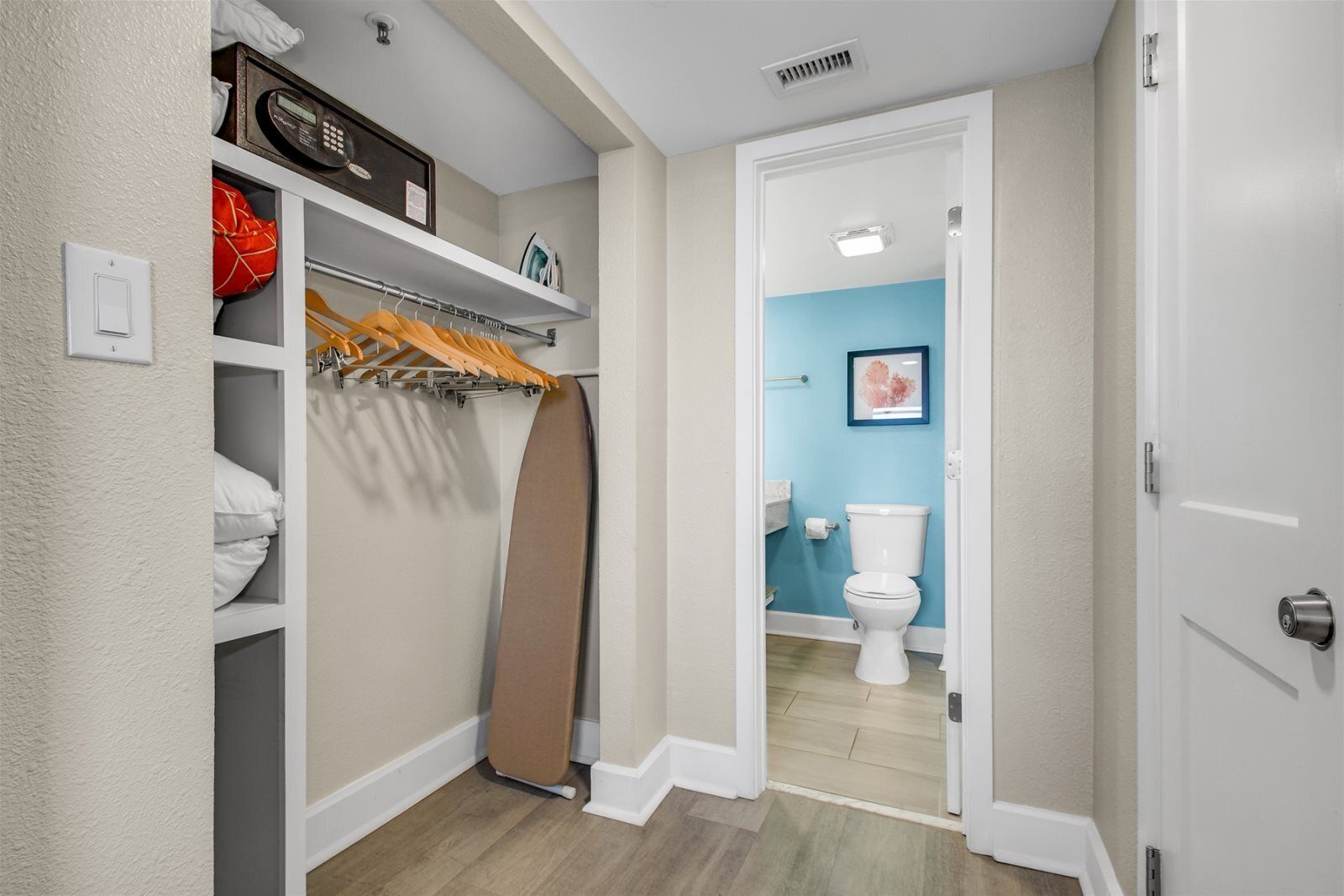 A hallway leading to a bathroom with a toilet and a blue wall. There is a white door on the right and a closet with a hanging rack and shelves on the left.