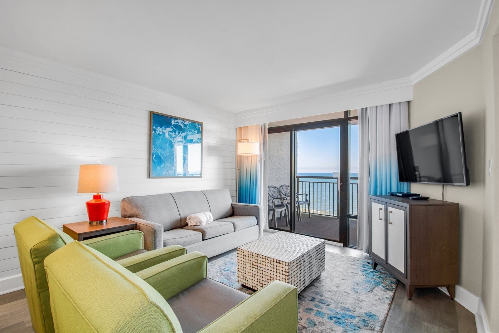 A hotel room with a grey sofa, green armchairs, a red lamp, a TV, and a balcony with a view of the sea.