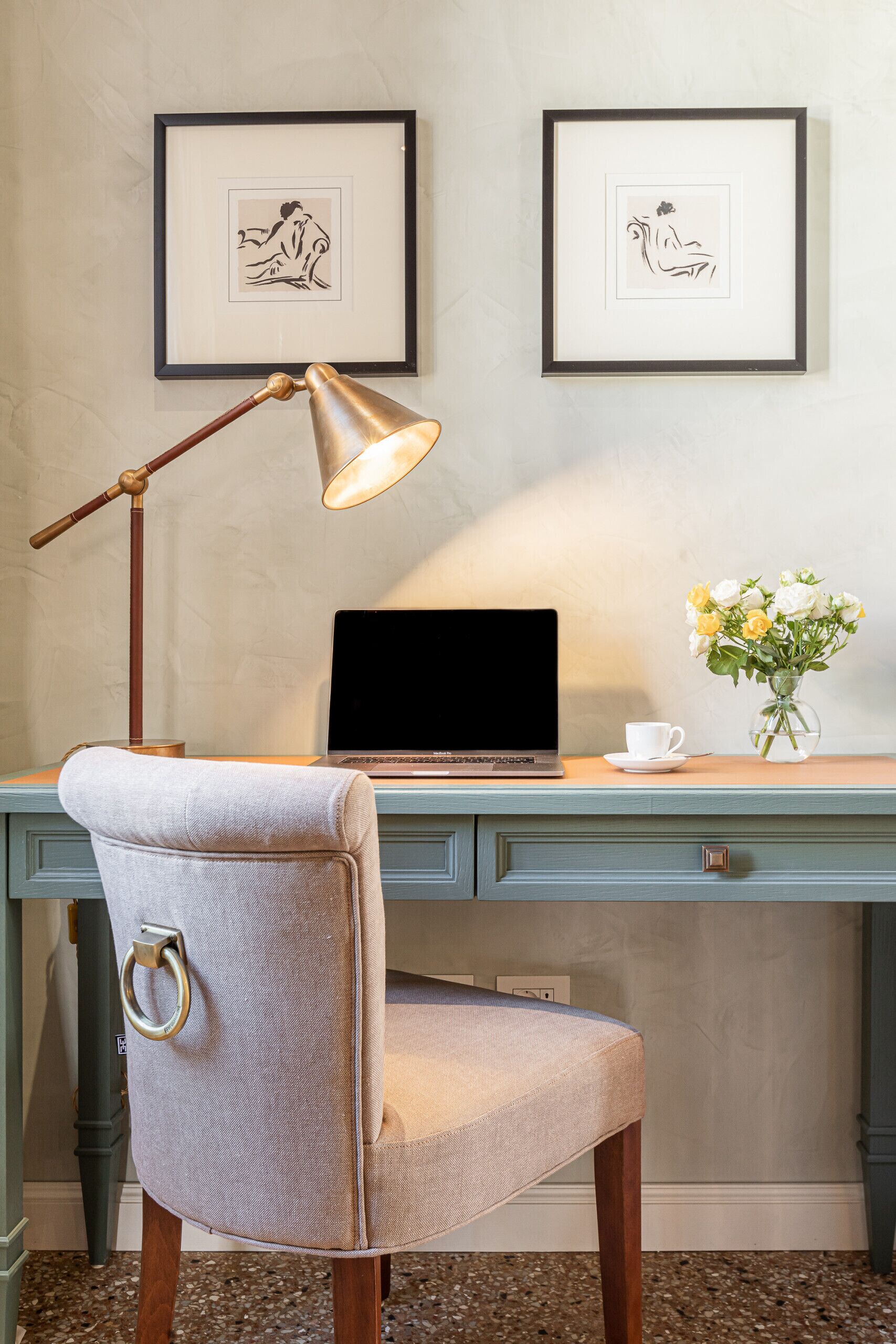A desk with a laptop, a lamp, a vase with flowers, and framed artwork on the wall.